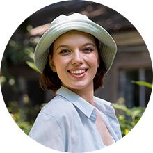 Smiling young woman wearing a light blue bucket hat and matching button-down shirt in a sunlit outdoor garden setting.