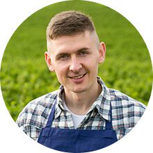 Smiling young professional in a blue apron and checkered shirt standing in a vibrant green agricultural field.
