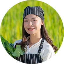A young female chef wearing a black and white striped pinstripe hat and apron, holding fresh leafy greens in an outdoor field setting.