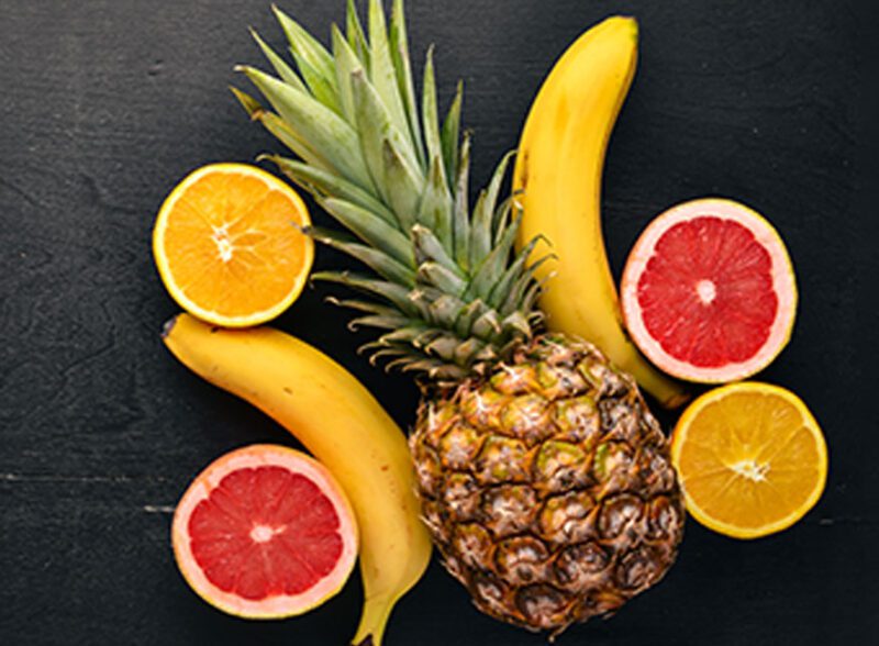 A top-down view of a whole pineapple, bananas, and sliced oranges and grapefruits arranged on a dark, textured background.