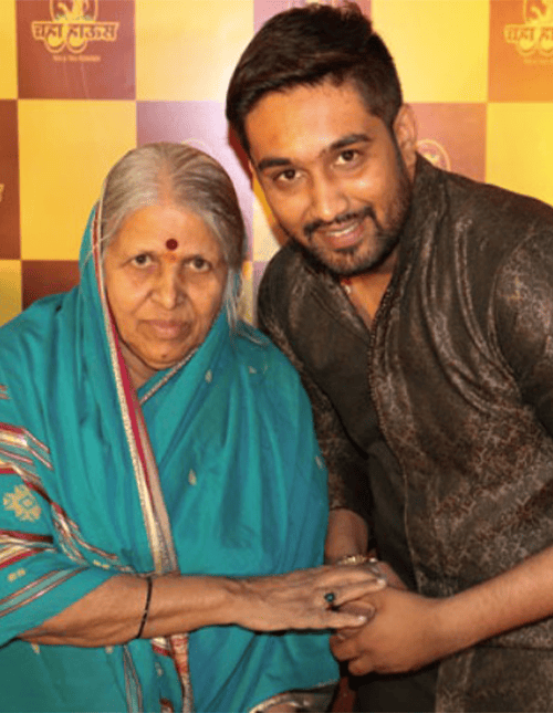 An elderly woman in a teal saree and a young man in a dark kurta posing together in front of a 'Chaha House' branded backdrop.